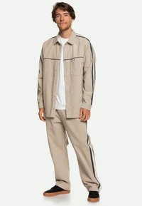 Camisa de algodão bege de botões com riscas pretas, combinada com calças a condizer. Apresenta um bolso no peito e um tecido texturizado. Ténis pretos.