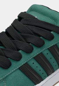 Πράσινο καστόρινο αθλητικό παπούτσι με μαύρα υφασμάτινα κορδόνια, τρεις μαύρες ρίγες, διακοσμητικές διατρήσεις και λευκή σόλα. Διαθέτει αντίθετα ραφτικά σχέδια.