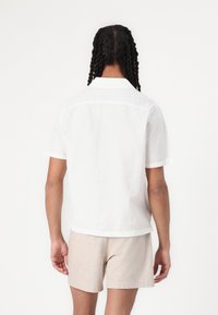 Chemise blanche à manches courtes avec un tissu texturé, présentant un motif à carreaux subtil et un col classique, assortie à un short beige clair.
