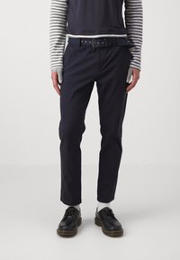 Pantalon marine à coupe droite, avec une ceinture en métal et des œillets. Associé à une chemise rayée et des chaussures noires.