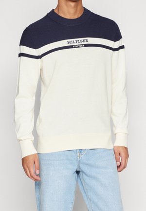 Man draagt een creme- en marineblauwe trui met lange mouwen met de tekst "Hilfiger New York" en lichtblauwe spijkerbroek, staand tegen een effen achtergrond.