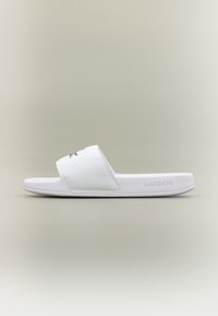 White slide sandal made of synthetic material, featuring a single wide strap with a green logo, flat sole, and minimalistic design.