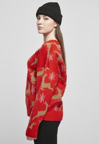 Czerwony sweter z dzianiny z wzorami reniferów i śnieżynek w kolorze złotym i białym. Modelka nosi czarną czapkę. Luźny krój, teksturowana tkanina.