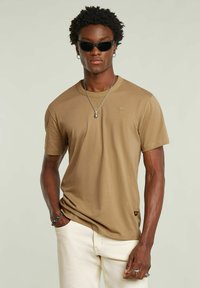 T-shirt marrone a manica corta realizzato in tessuto morbido. Presenta un colletto a girocollo ribassato e un logo discreto vicino al petto. Abbinato a pantaloni bianchi.