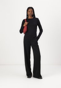 Haut noir à manches longues avec un col rond, associé à un pantalon noir ample. Un petit sac bandoulière rouge est tenu sur le côté.