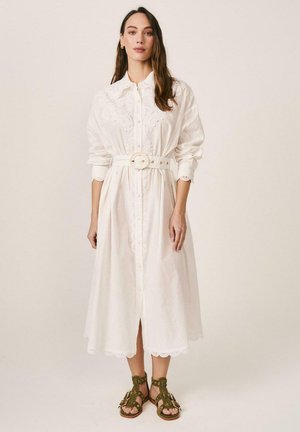 Mujer con un vestido midi blanco bordado y un cinturón a juego, y sandalias de tiras en verde oliva, posando frente a un fondo liso.