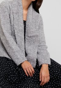 Grå, fluffig cardigan med öppen front, strukturerad tyg och långa ärmar. Bärs över en svart klänning med vita prickar.