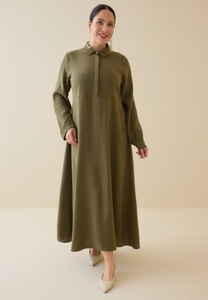 Kvinde iført en lang olivengrøn kjole med krave og lange ærmer, iført beige spidse sko og store runde øreringe mod en ensfarvet beige baggrund.