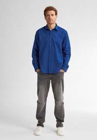 Košile s dlouhým rukávem, modrá, z měkké bavlny, rovného střihu, spárovaná se světle šedými džíny a bílými teniskami, stojící na neutrálním pozadí.