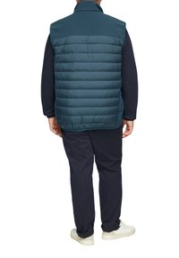 Tannengrüner Puffer-Weste mit hohem Kragen, die eine gesteppten Textur und ein glattes Rückenteil aufweist, kombiniert mit marineblauen Hosen und weißen Sneakers.