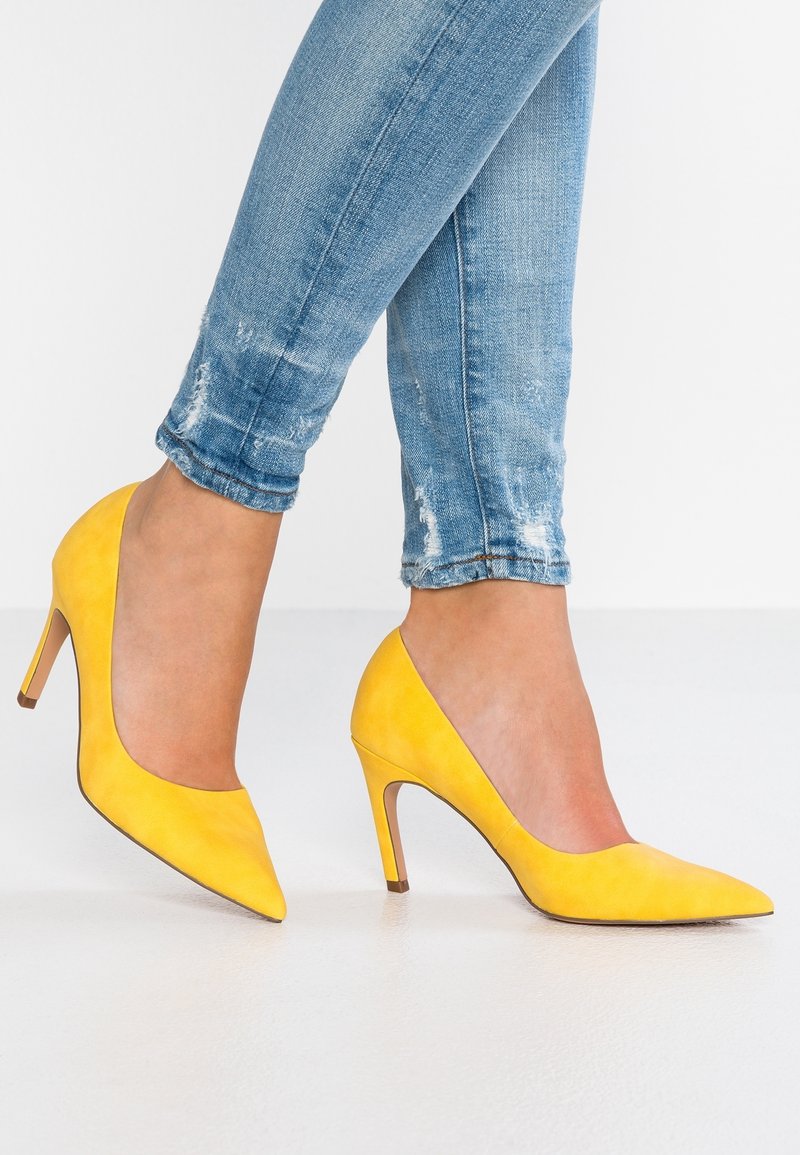 Escarpins à talons hauts en daim jaune avec un bout pointu et un talon fin, associés à un jean skinny bleu déchiré, sur une surface blanche.