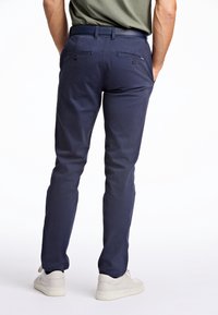 Mężczyzna w granatowych spodniach, białych trampkach i oliwkowej koszuli, stojący z rękami w tylnych kieszeniach na białym tle.