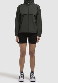 Mörkgrön vattentät jacka med dragkedja fram, stor bröstficka och sidodragkedjor, kombinerad med svarta shorts och högstövlar.