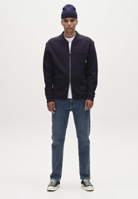 Mornarško jakna iz volnenega mešanega materiala z zadrgo in sprednjim žepom, v kombinaciji s svetlo modrimi kavbojkami in črno-belimi supergami. Manekenka nosi kapo.