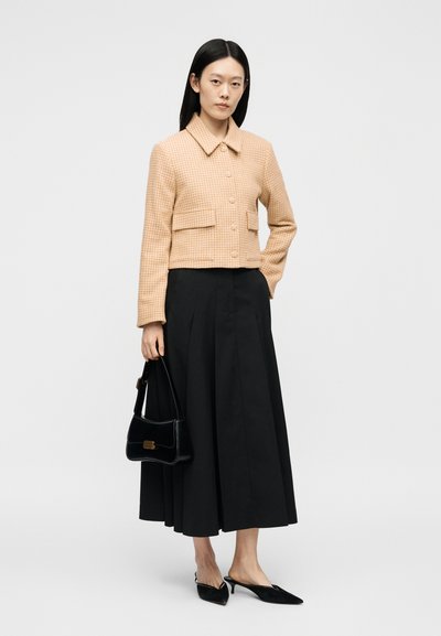 Femme portant une veste beige à carreaux boutonnée et une jupe midi noire, tenant un sac à main noir et portant des escarpins noirs à bout pointu et talons bas.