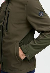 Grüne Softshelljacke mit hohem Kragen, Reißverschluss-Brusttasche, Seitentaschen und einem Logopatch am linken Ärmel. Glatte Textur.