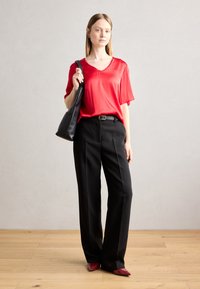 Blusa rossa in satin con maniche corte, pantaloni neri sartoriali e una borsa a tracolla nera. Il modello indossa ballerine rosse a punta, in piedi su un pavimento di legno.
