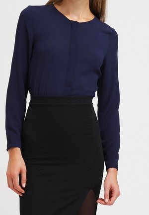 Marineblaue Bluse mit langen Ärmeln und rundem Ausschnitt, kombiniert mit einem schwarzen Bleistiftrock mit strukturiertem Muster und Oberschenkelschlitz.