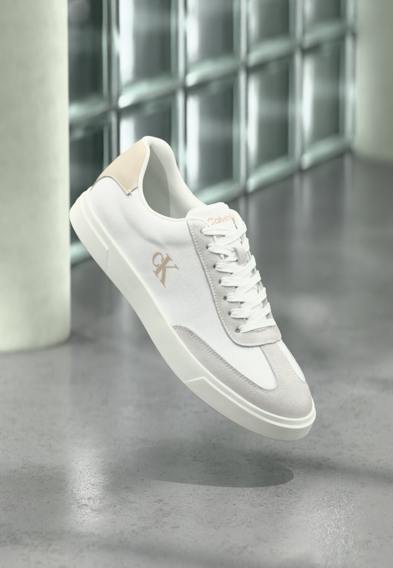 Baskets blanches Calvin Klein avec accents en daim beige et logo, flottant au-dessus d'un sol gris lisse avec un mur en blocs de verre en arrière-plan.
