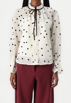 Blouse blanche transparente à pois noirs, col montant avec lien en ruban noir, boutons sur le devant, portée avec un pantalon bordeaux taille haute à jambes larges.