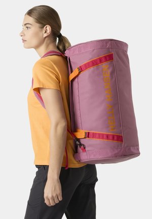 Mujer con cabello castaño claro recogido en una coleta, vestida con una camiseta naranja, lleva una gran bolsa cilíndrica rosa de Helly Hansen en la espalda.