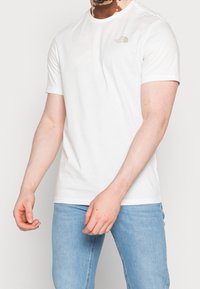 Muž v bílém tričku s krátkým rukávem značky The North Face a světle modrých džínech, stojící na pozadí v jednotné světlé barvě.