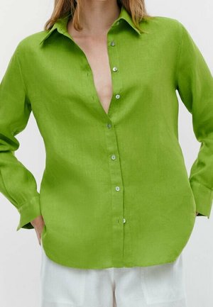 Femme portant une chemise boutonnée vert vif avec les premiers boutons déboutonnés et un pantalon blanc, les mains dans les poches.