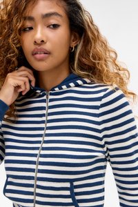 Jonge vrouw met krullend haar, die gouden ringoorbellen en een blauw-wit gestreepte hoodie met ritssluiting draagt, terwijl ze de capuchon bij haar nek aanpast.