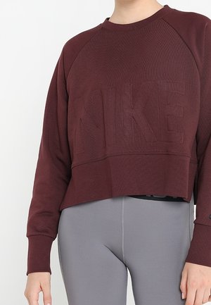 Osoba ubrana w ciemnoczerwony krótki sweter Nike i jasnoszare dopasowane legginsy na jednolitym białym tle.