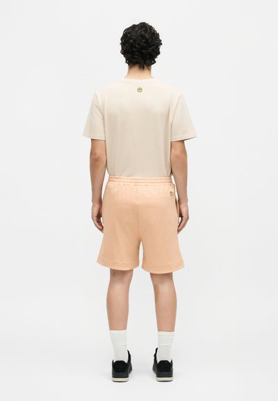 Άνδρας με σγουρά σκούρα μαλλιά που φοράει μπεζ μπλουζάκι και ανοιχτόχρωμα ροδακινί σορτς, στέκεται με την πλάτη σε απλό λευκό φόντο.