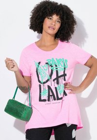 Roze T-shirt met korte mouwen met graffiti-achtige tekst. Het model houdt een groene, gestructureerde handtas vast, gecombineerd met donkere jeans versierd met harten.