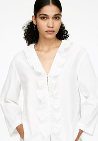White button-up shirt featuring a ruffled neckline and front placket, with long sleeves and a relaxed fit, made from a lightweight fabric.