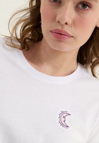 Noor naine laineliste pruuni juustega, kes kannab valget särki, millel on kaeluse lähedal väike lilla poolkuu näokujutisega.