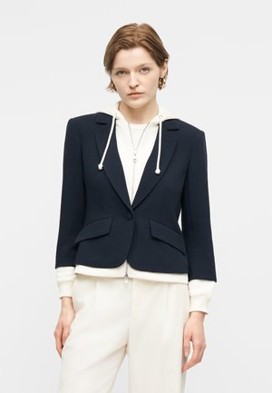 Γυναίκα με κοντά μαλλιά που φορά χρυσούς κρίκους σκουλαρίκια, λευκό φούτερ με φερμουάρ κάτω από ένα εφαρμοστό μαύρο σακάκι, και φαρδιά λευκά παντελόνια μπροστά σε απλό φόντο.