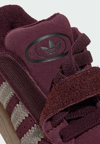 Gros plan d'une basket en daim bordeaux avec trois bandes grises, logo Adidas noir sur la languette et une bride en velcro sur des lacets assortis.