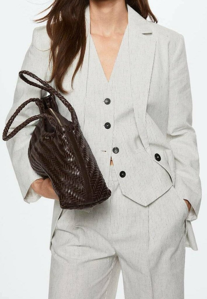 Femme portant un costume clair à fines rayures tenant un grand sac à main en cuir tressé brun foncé avec des poignées tressées.