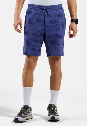 Lila Shorts mit einem Kordelzugbund, die ein Muster aus Wäldern und Wildtieren in dunkleren Farbtönen aufweisen. Kombiniert mit grauen Sneakers und weißen Socken.