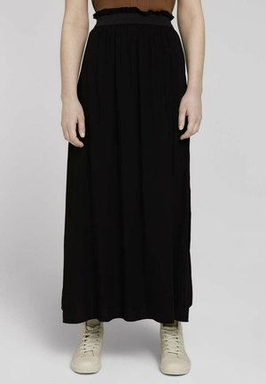 Femme portant une jupe maxi noire taille haute associée à des baskets montantes beiges sur un fond uni.