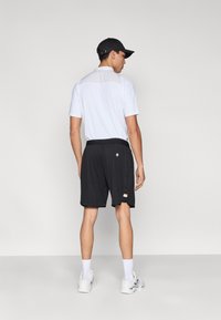 Bela majica s kratkimi rokavi in teksturirano hrbtno stranjo, črne športne kratke hlače z diskretnim logotipom ter bele nogavice, ki so združene s sivimi supergami.