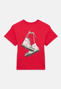 Κόκκινο μπλουζάκι με κοντά μανίκια που απεικονίζει ένα γραφικό με ροζ αθλητικά παπούτσια να κρέμονται. Η λέξη «αγάπη» είναι σχεδιασμένη πάνω από τα παπούτσια με λευκό χρώμα.