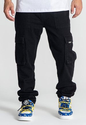 Pantalones cargo negros con bolsillos laterales, dobladillos ajustados y un corte holgado. Llevados con zapatillas azules con cordones amarillos y patrones gráficos.