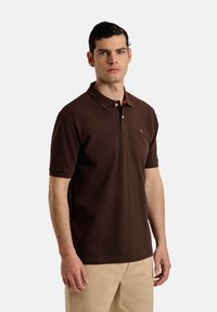 Camisa polo marrón de algodón, con cuello, mangas cortas y un detalle naranja en el cuello. Incluye un pequeño logo en el pecho.