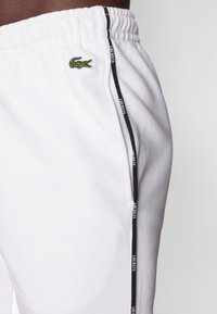 Vita bomullsjoggers med en texturerad yta, med en grön krokodil-logotyp och svarta sidostråk med "LACOSTE"-märkning.