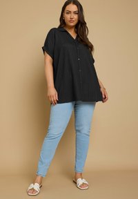 Camicetta nera con bottoni, in tessuto a coste e maniche corte arrotolate, abbinata a jeans skinny azzurri e sandali bianchi in stile scivolo.