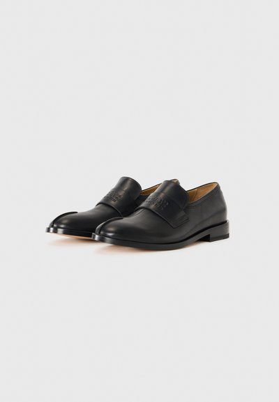 Des chaussures en cuir noir présentent un bout arrondi, une texture lisse et une sangle proéminente sur le dessus avec des détails en relief. Talon bas.
