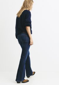 Temno modri flare jeans z rahlim bledanjem, s srednjim pasom, zadnjimi žepki in gladko teksturo, v kombinaciji z rjavimi gležnjači.