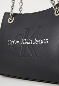 Borsa a mano in pelle nera con logo "CK" in rilievo e il testo "Calvin Klein Jeans", con tracolla in catena argentata e dettagli a occhiello.