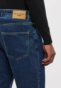 Dark blue denim jeans feature a leather label with "Marc O'Polo DENIM" at the back waistband, visible stitching, and a standard pocket design.