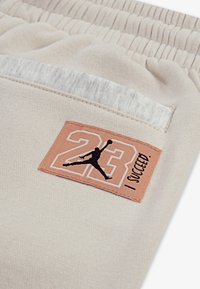 Bež joggeri s teksturiranim materijalom, elastičnim pojasom i istaknutom narančastom označenom etiketom s "23" i grafikom crnog silueta.