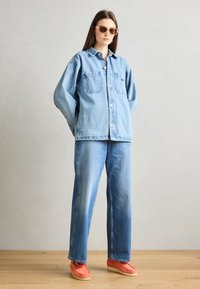 Camisa de mezclilla oversize con dos bolsillos en el pecho, combinada con pantalones vaqueros anchos de color azul claro y zapatillas naranjas brillantes. Gafas de sol marrones.